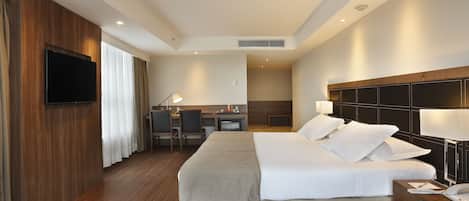 A hotel room with a large bed, bedside table with a lamp, a desk with a chair, a TV mounted on the wall, and a window with curtains.