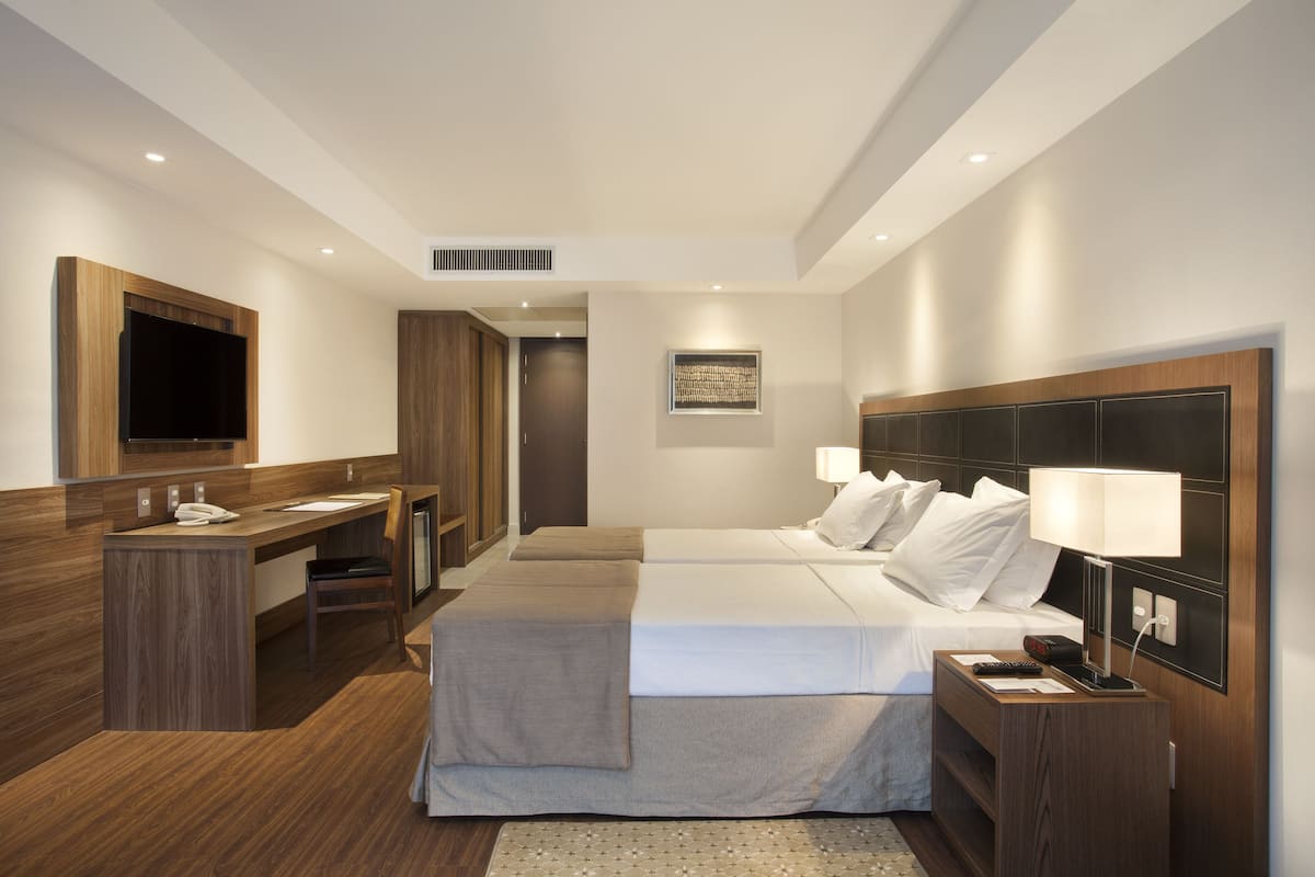 Una habitación de hotel moderna con un escritorio de madera, dos camas, un televisor y una mesita de noche con una lámpara.