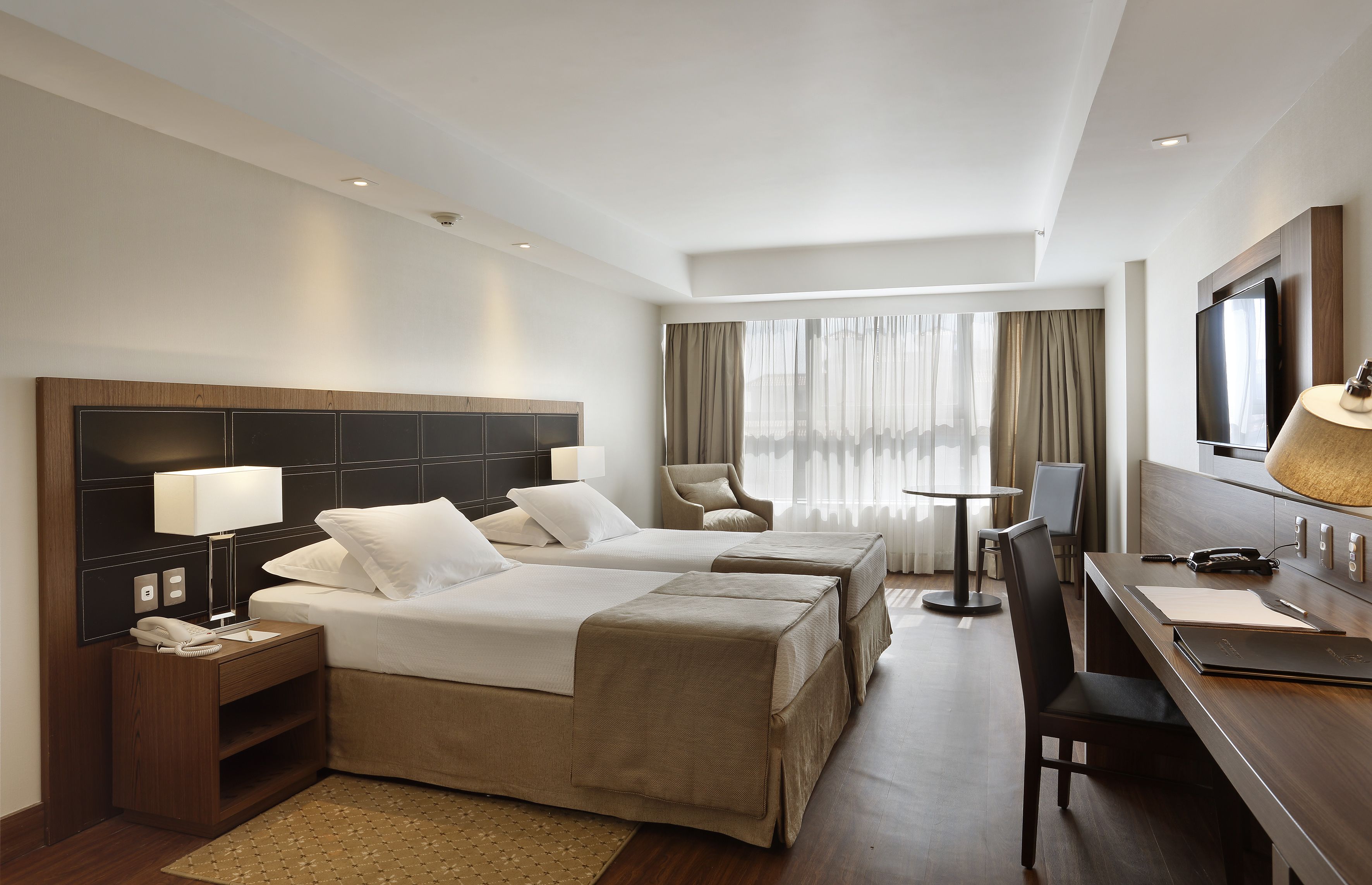 A hotel room with two beds, a desk, a chair, a TV, and a window with curtains.
