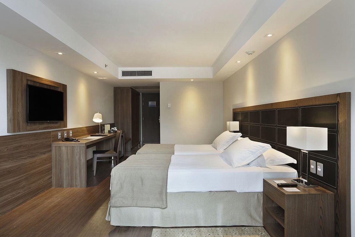 A hotel room with a large bed, a desk, a chair, a TV mounted on the wall, and a wooden headboard.