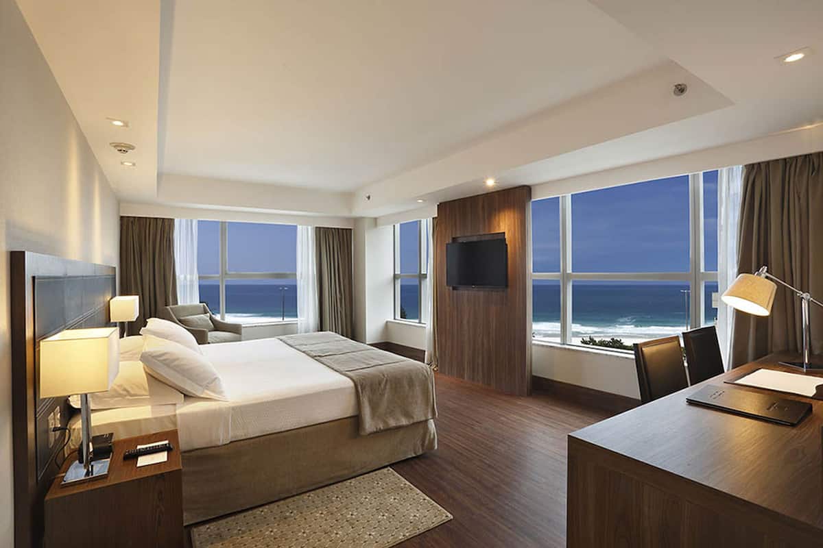 Habitación de hotel moderna con una cama grande, un escritorio y un televisor. La habitación tiene ventanas amplias con vista al océano.