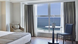 A hotel room with a large bed, a chair, a small table with a plant, and a view of the ocean through a large window. - Windsor Oceanico (Rio de Janeiro)