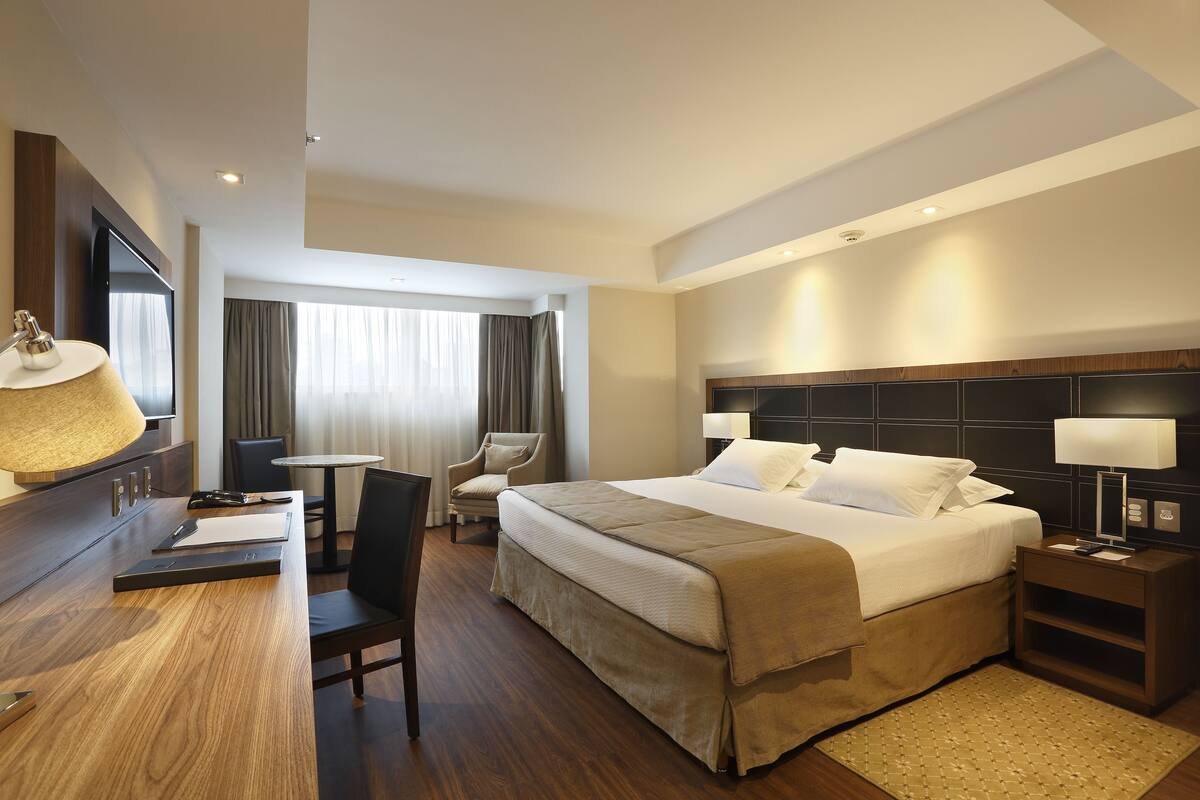 Una habitación de hotel con cama, escritorio, silla, televisión y ventana con cortinas.