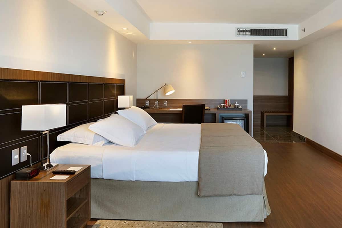 Habitación de hotel con una cama grande, mesitas de noche, un escritorio, una silla, un televisor montado en la pared y un baño visible a través de una puerta abierta.