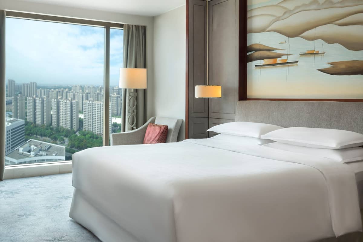 A hotel room with a large bed, a view of the city, and a painting on the wall.