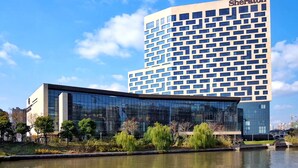 A Sheraton hotel with a modern glass facade, a courtyard with trees, and a body of water in front. - Sheraton Shanghai Jiading Hotel (Shanghai)