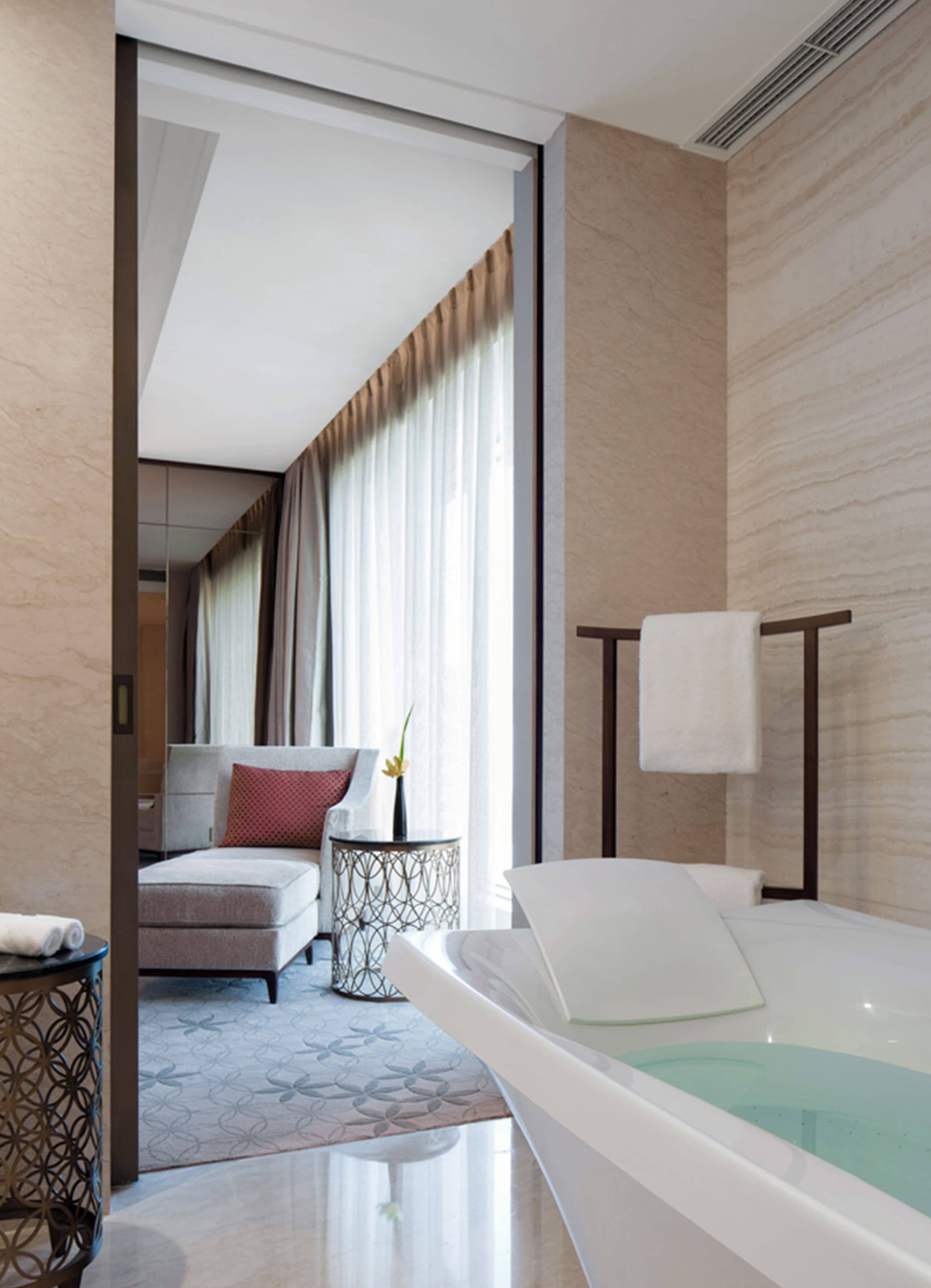 A modern hotel room with a large bathtub, a sofa, a side table with a vase, and a view of the hallway.
