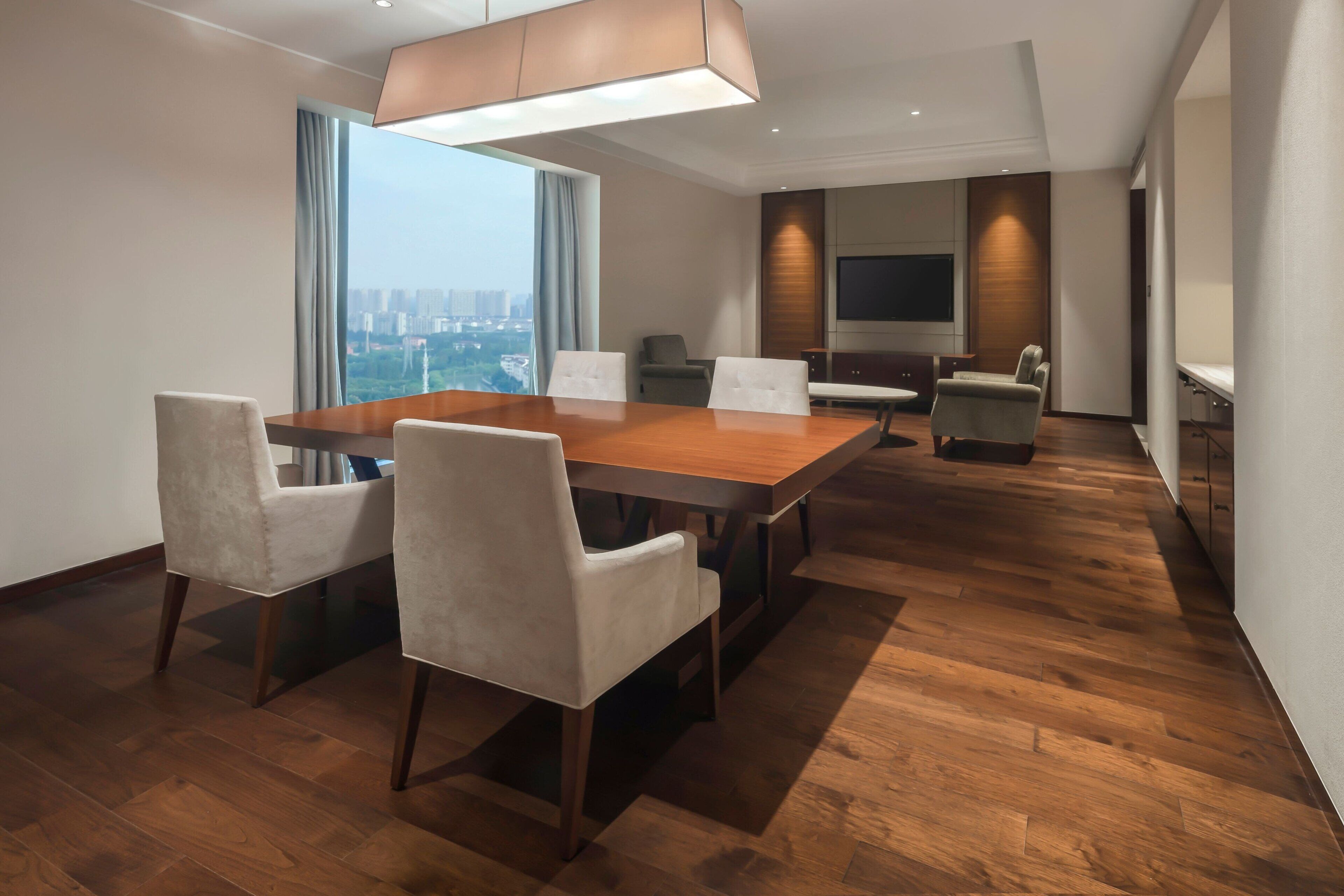 A modern hotel room with a large wooden dining table, white upholstered chairs, a flat-screen TV mounted on the wall, and a city view through large windows.