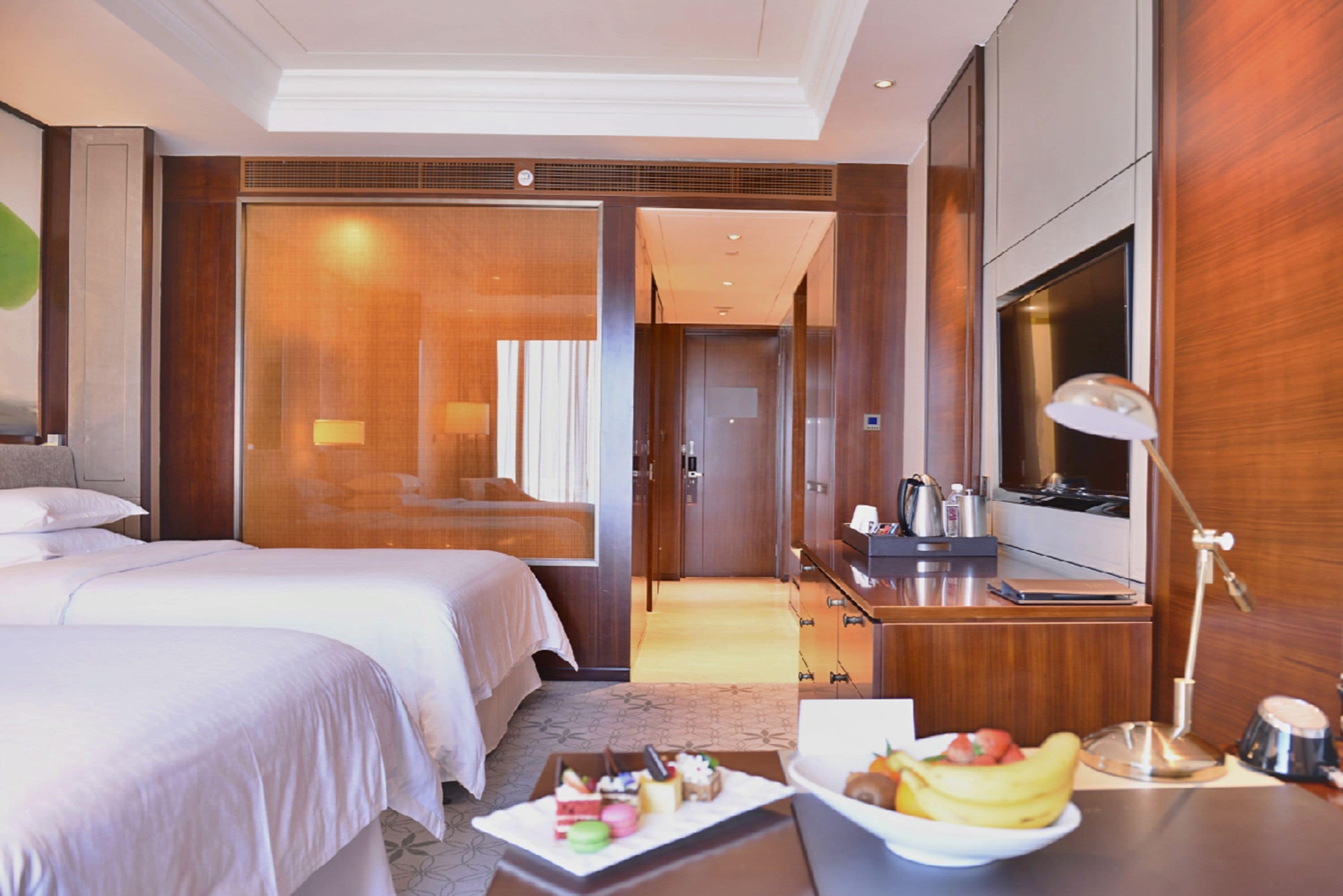 Habitación de hotel con dos camas, un escritorio con lámpara, televisor y una bandeja de desayuno con fruta y pastelería.