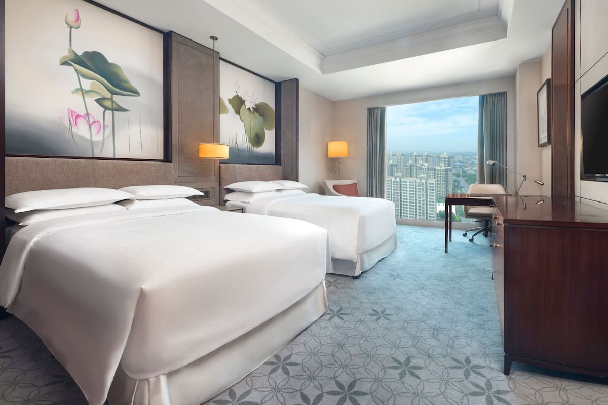 A hotel room with two beds, a desk, a TV, and large windows with a city view.