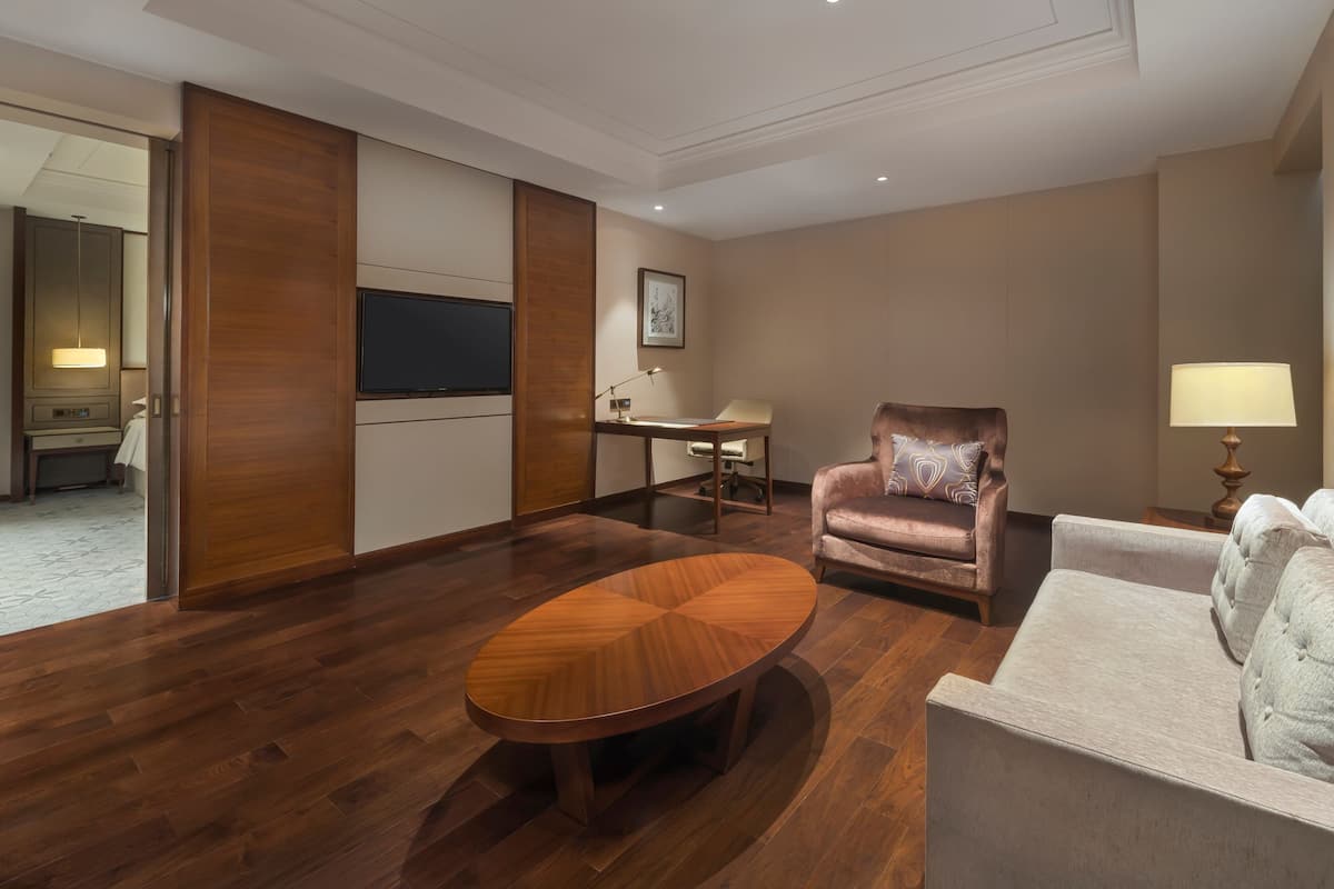 A modern hotel room with a wooden floor, a round coffee table, a sofa, a desk with a lamp, and a television.