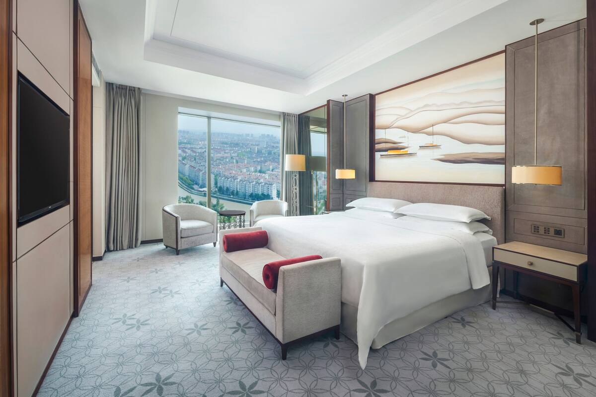 A hotel room with a large bed, a TV, a desk, and a view of the city.