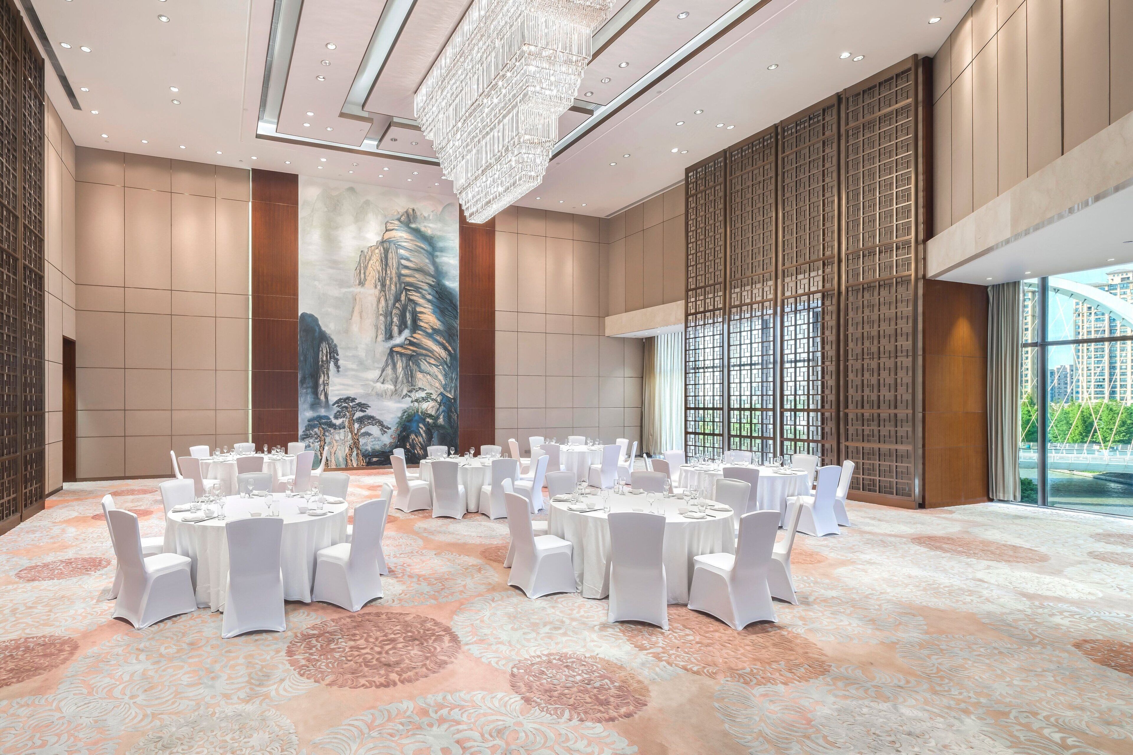 Une vaste salle de banquet, avec des tables rondes dressées pour le repas, un grand lustre et une fresque murale.