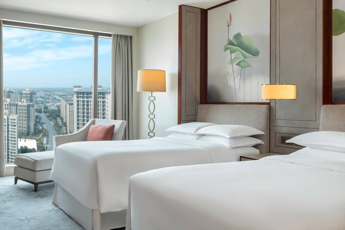 A hotel room with two beds, a desk, and a chair. There is a large window with a view of a cityscape.