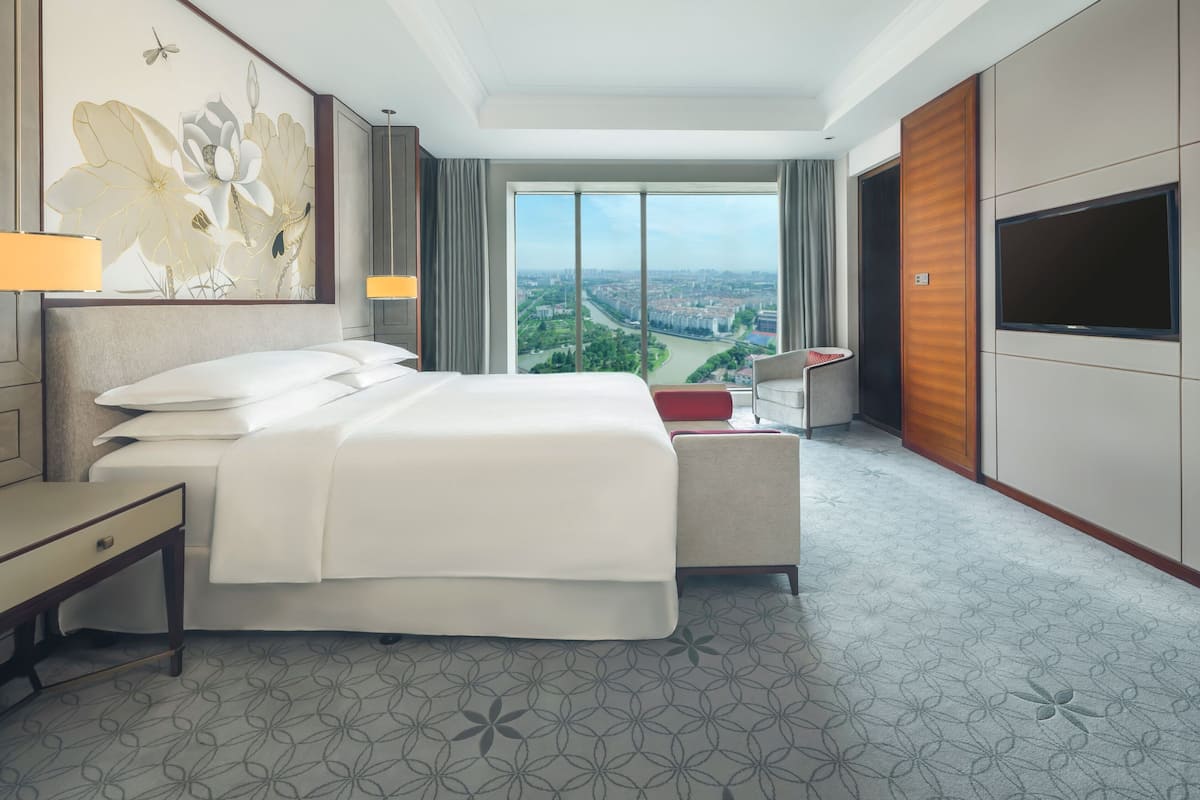 A hotel room with a large bed, a desk, a chair, a TV, and a view of a cityscape.