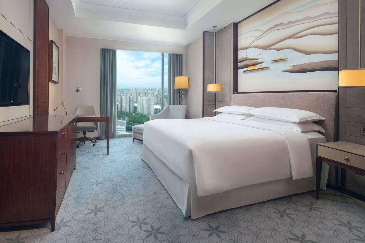 A hotel room with a large bed, a desk, a chair, and a view of the city.