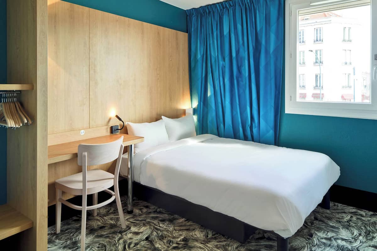 A hotel room with a bed, a desk, a chair, and a view of buildings through a window.