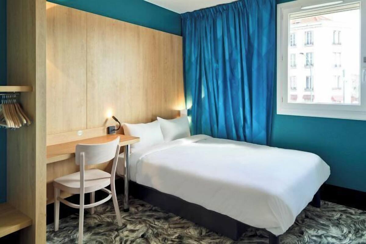 A hotel room with a bed, a desk, and a chair. There is a window with blue curtains and a view of buildings outside.