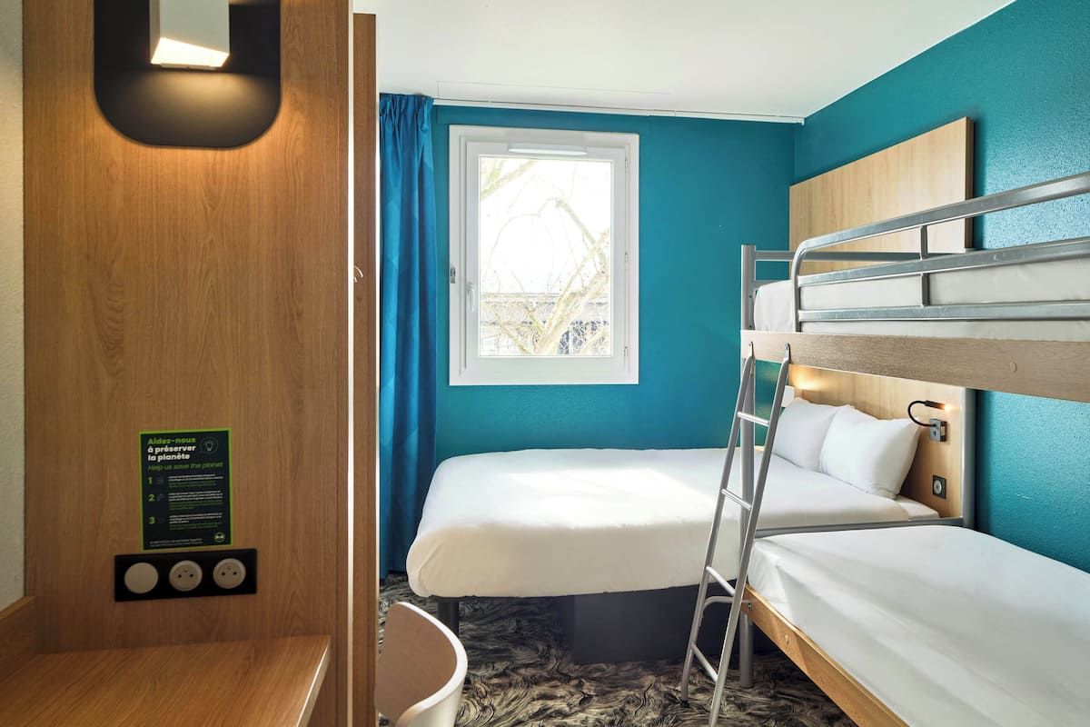 A compact hotel room with two bunk beds, a window with curtains, a wooden wall with a light fixture, and a sign about environmental preservation.
