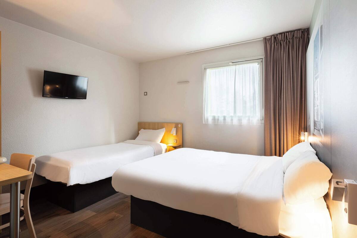 Une chambre d’hôtel avec deux lits simples, un téléviseur fixé au mur, un bureau et une fenêtre avec des rideaux.