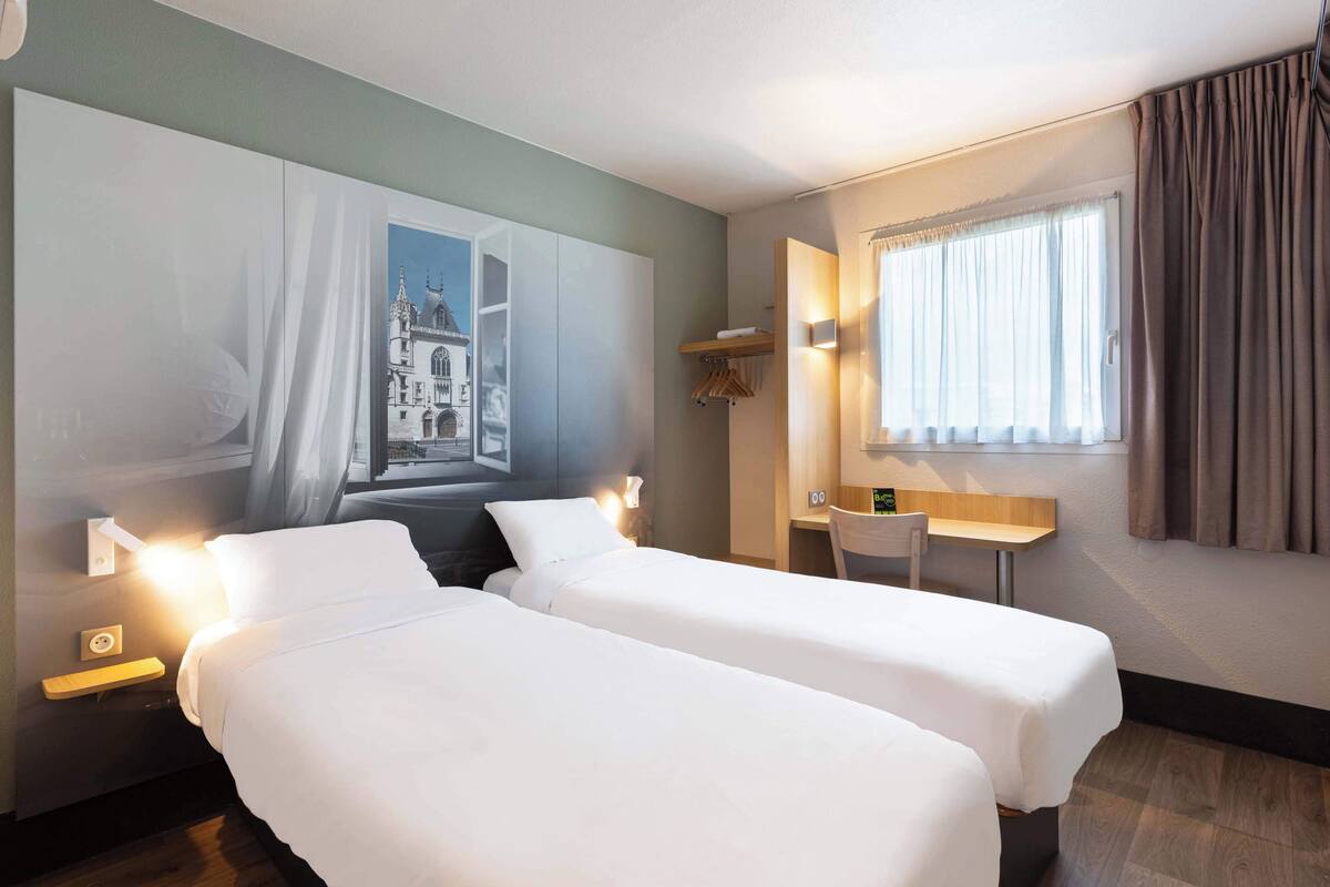 Une chambre d’hôtel avec deux lits, un bureau, une chaise, un tableau au mur et une fenêtre avec des rideaux.