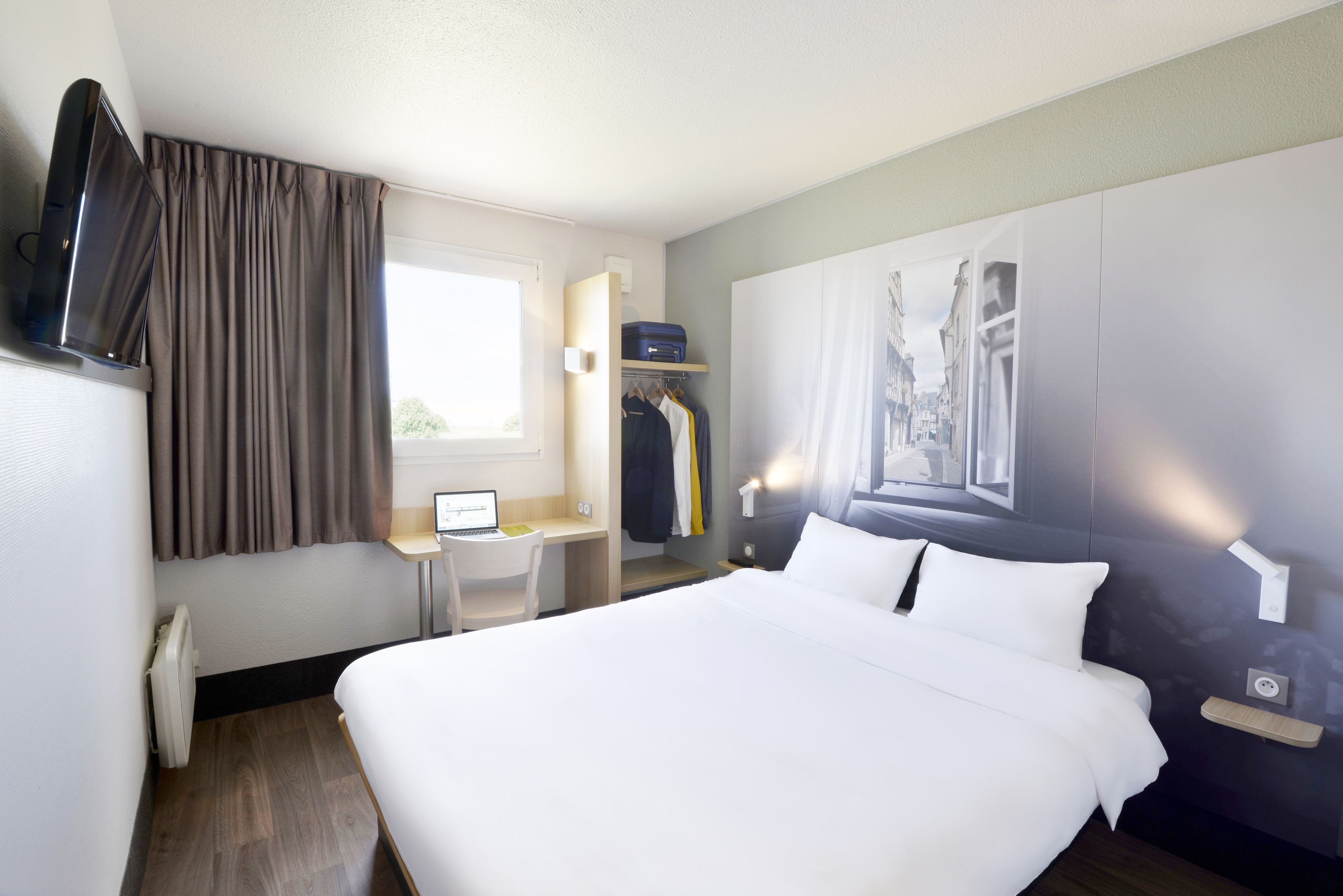 Habitació d'hotel amb un llit, un escriptori amb un ordinador, una televisió, una perxa de roba i una finestra amb cortines.