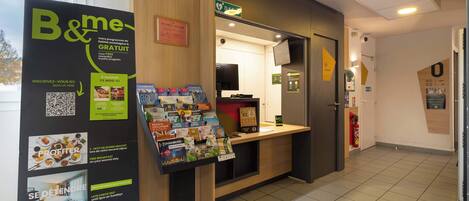 A hotel reception area with a reception desk, a sign for 'B&me' program, and informational brochures.