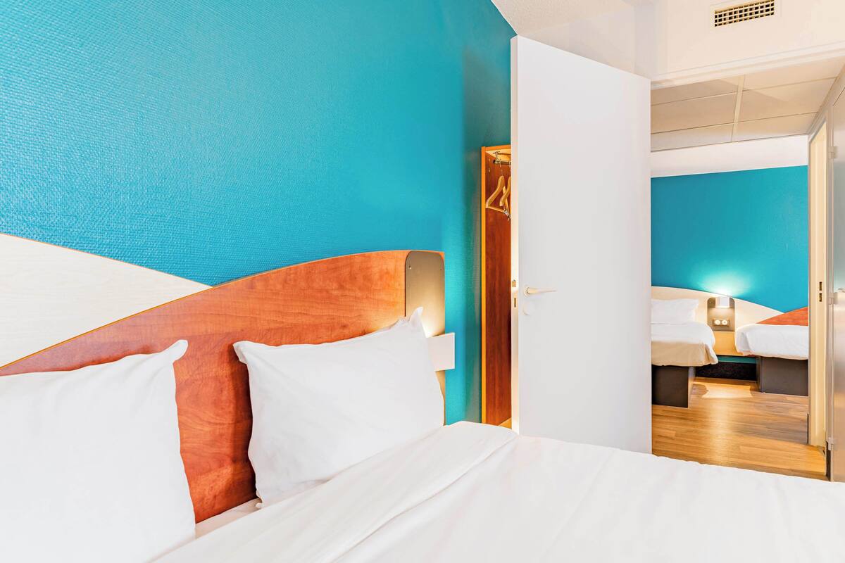 Habitación de hotel con una cama grande, cabecera de madera, pared de acento color verde azulado y un espejo que refleja otra habitación.