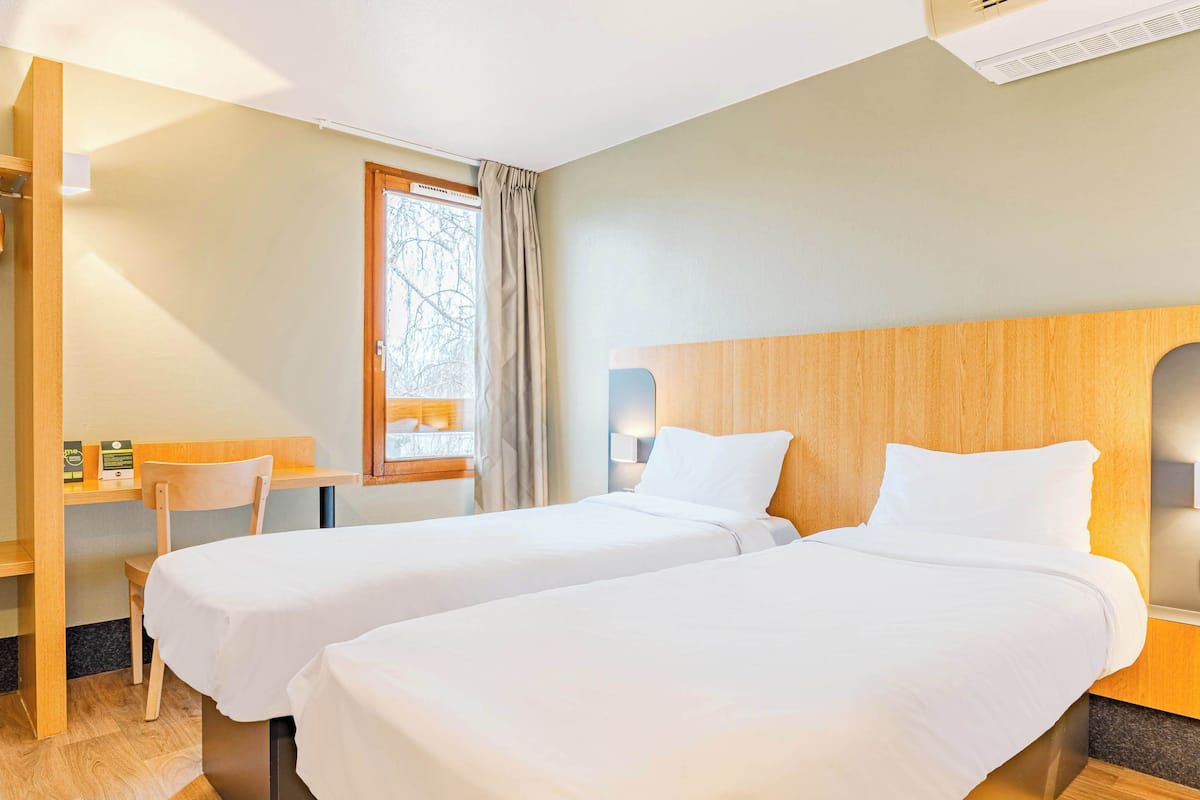 Habitación de hotel con dos camas, un escritorio, una silla, un ventanal con cortinas y un aire acondicionado de pared.