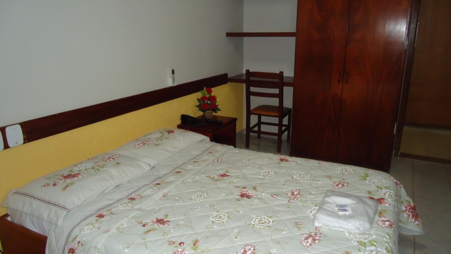 A bedroom with a bed, a chair, a wardrobe, and a nightstand with a vase of flowers.