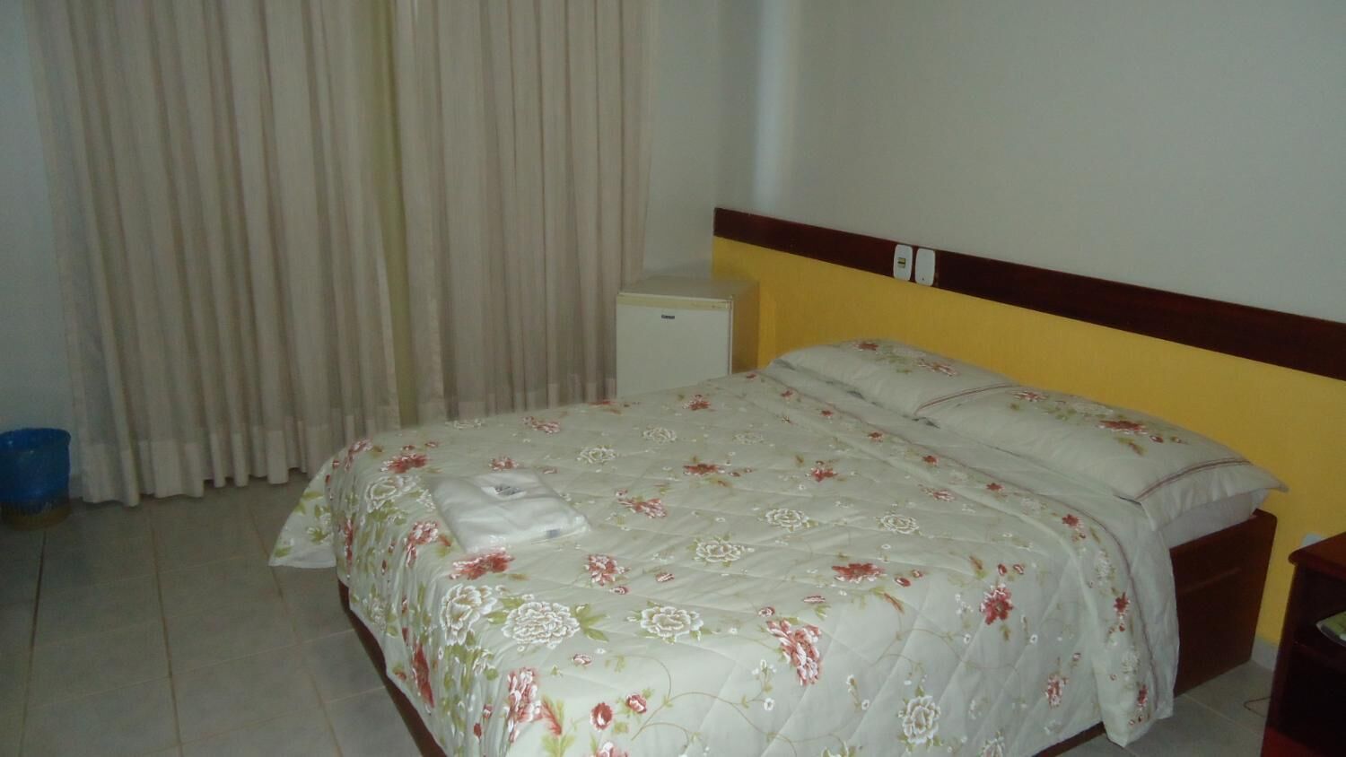 A bedroom with a bed, a small bedside table, a nightstand, a wall-mounted light switch, and a window with curtains.
