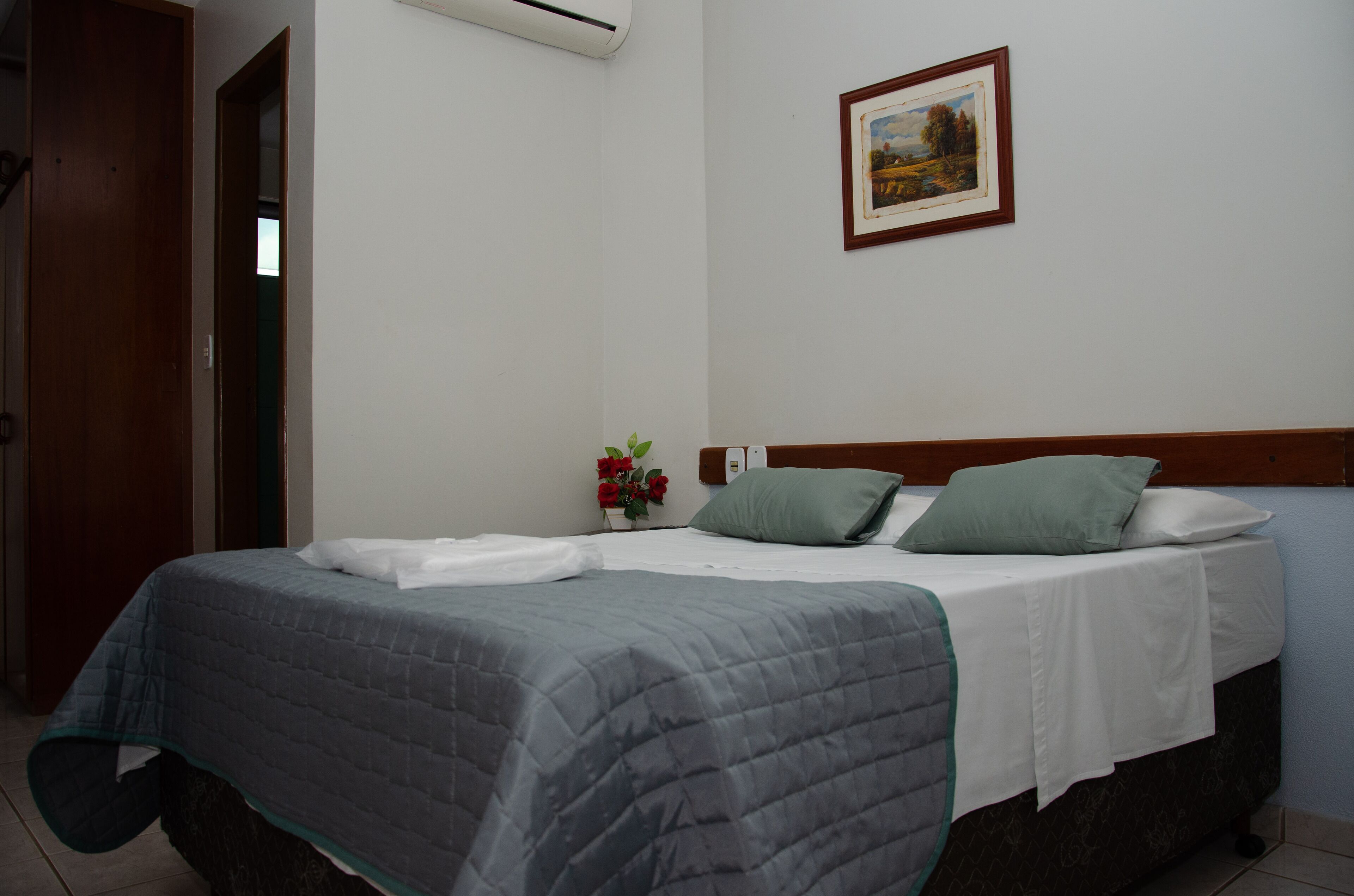 Una cama bien hecha con colcha gris, sábana blanca y dos almohadas. Encima de la cama, hay un cuadro en la pared.