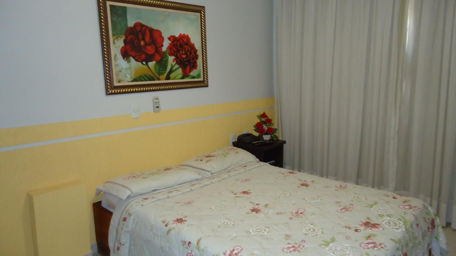 A room with a bed, a painting of flowers on the wall, a nightstand with a lamp, and a window with curtains.