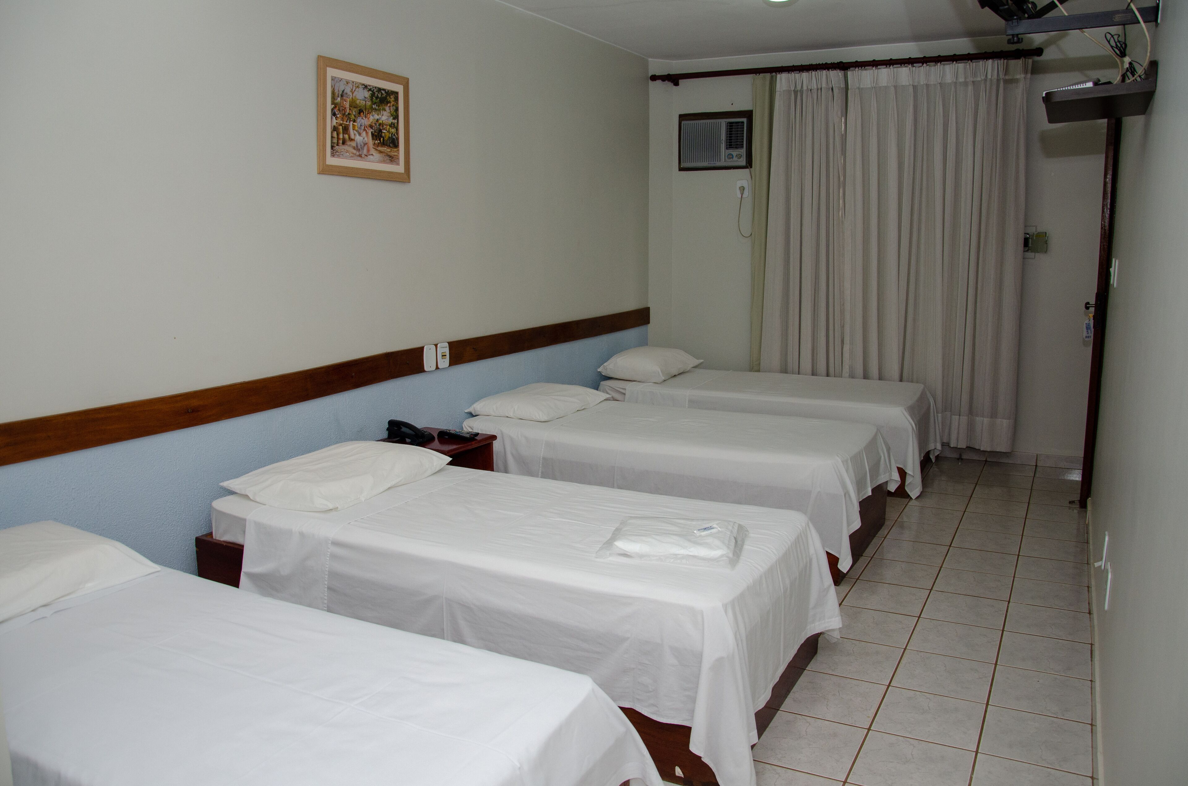 A hotel room with four single beds, a wooden headboard, a small bedside table with a phone, a picture on the wall, and a window with curtains.