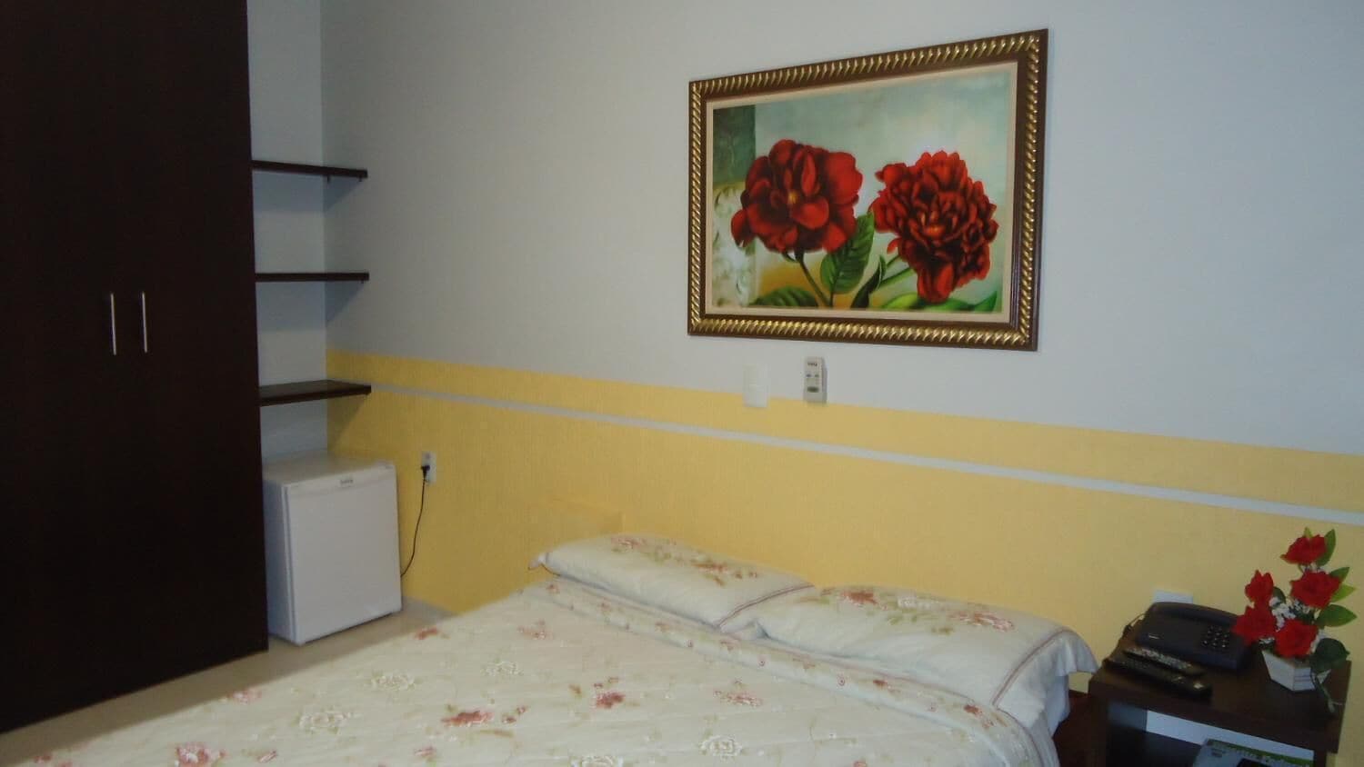 Une chambre à coucher avec un lit, un petit appareil électroménager blanc, un tableau représentant des fleurs rouges et un téléphone sur une table de chevet.