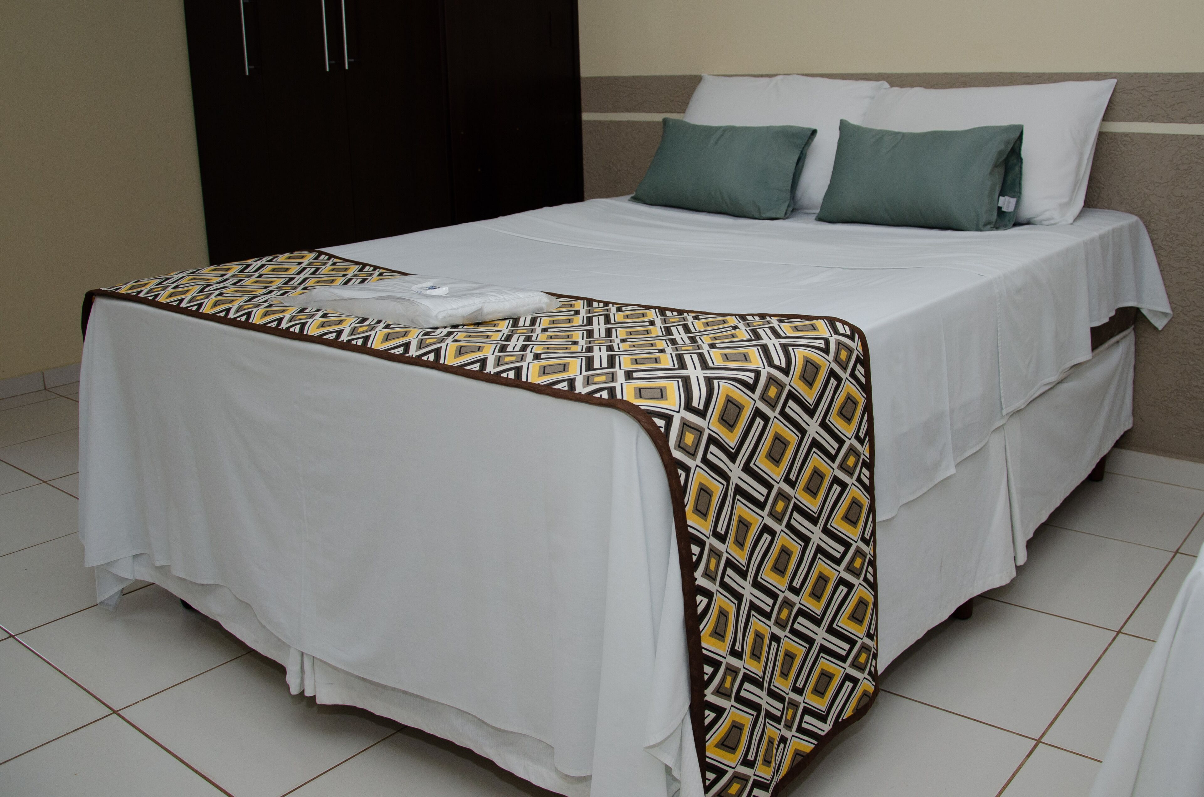 A neatly made bed with a patterned bedspread, a dark wardrobe, and a plain wall.