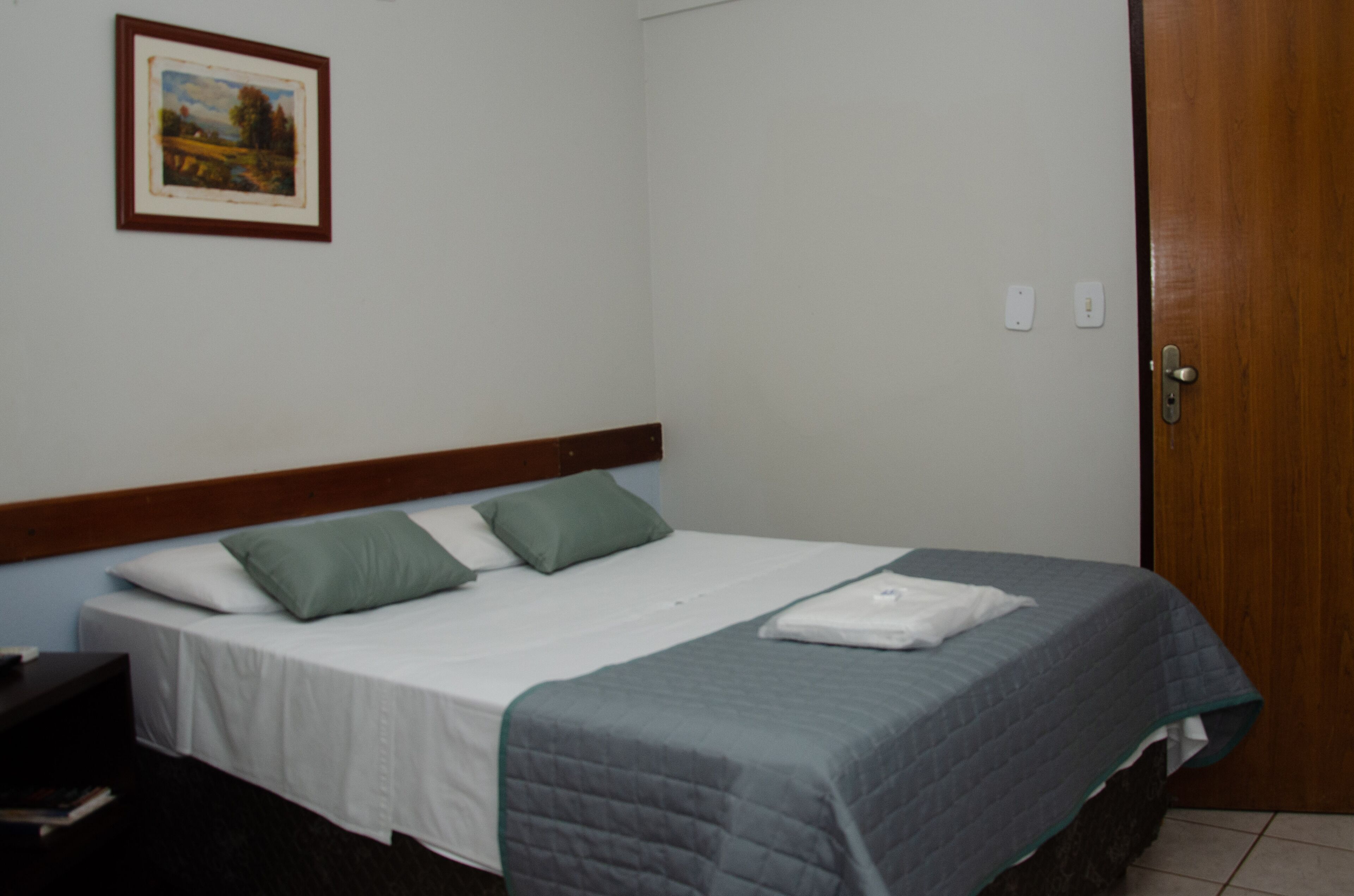 A neatly made bed with a grey quilt, two green pillows, and a white blanket folded at the foot.