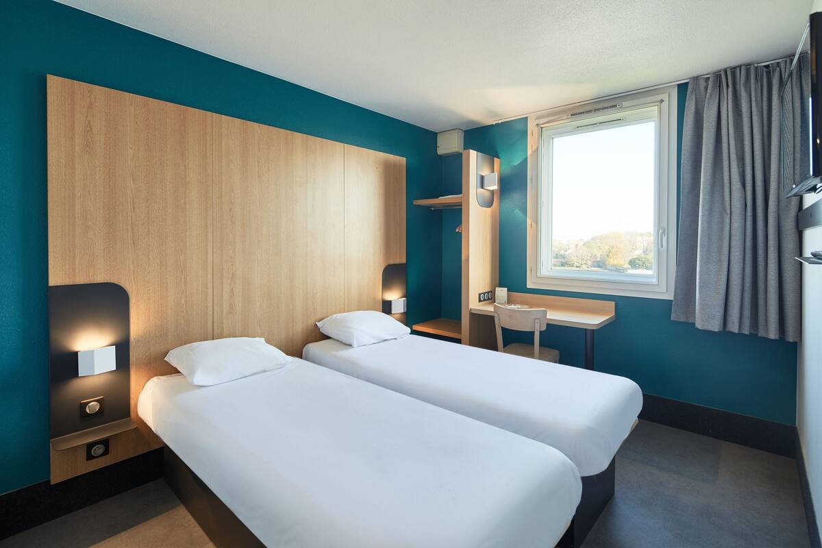 A hotel room with two single beds, a desk, a chair, a window with curtains, and a wooden headboard.
