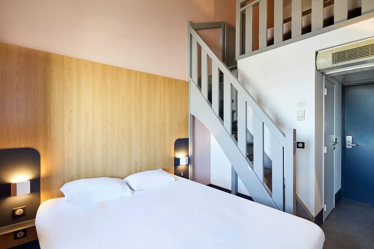 A hotel room with a bed, two bedside lamps, a wooden headboard, a staircase, and a door.