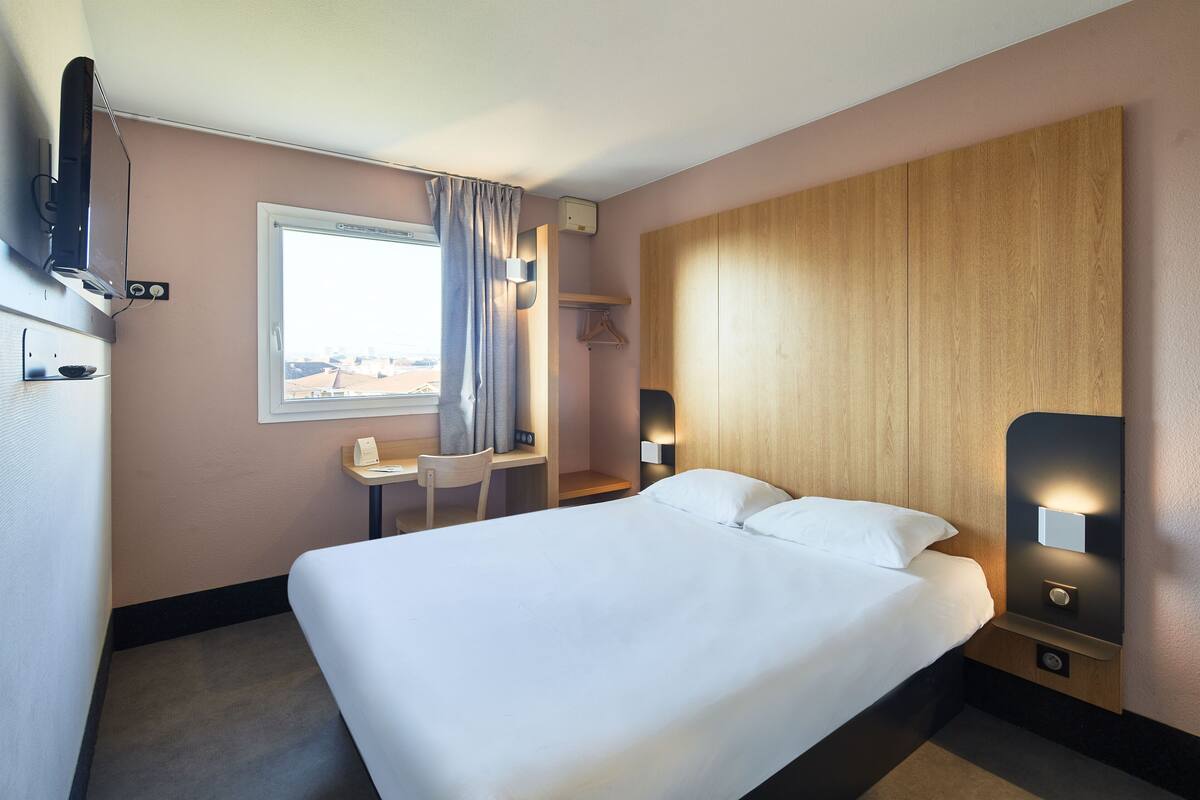 A hotel room with a large bed, a desk, a chair, a TV, and a window with curtains.