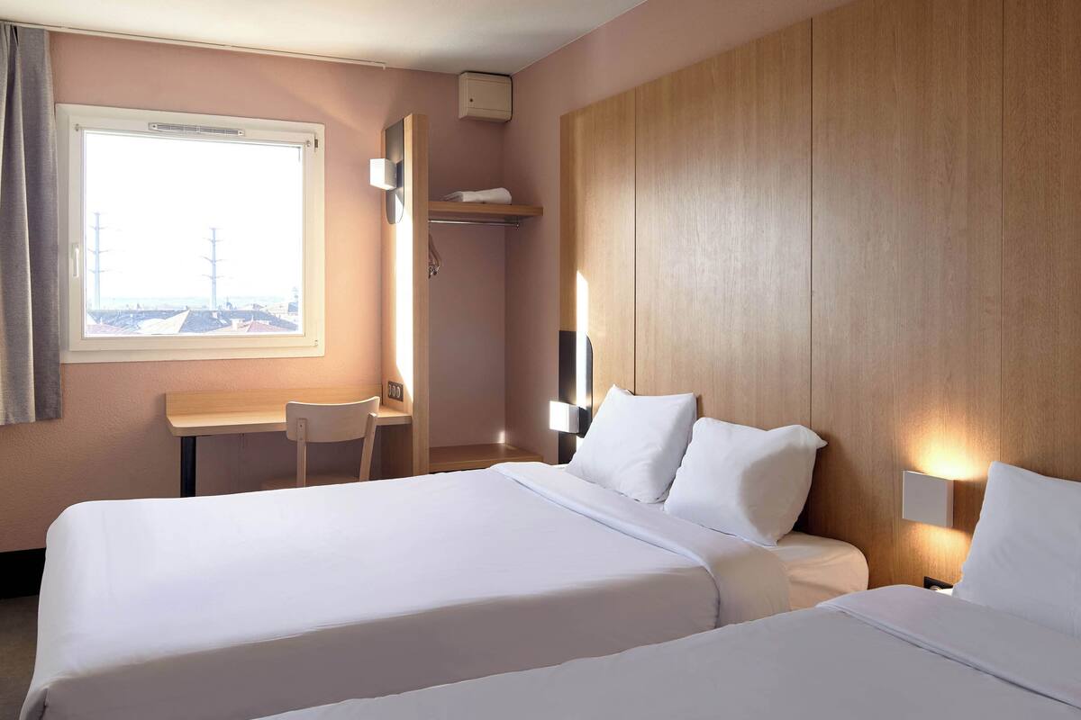 A hotel room with two beds, a desk, and a window with a view.