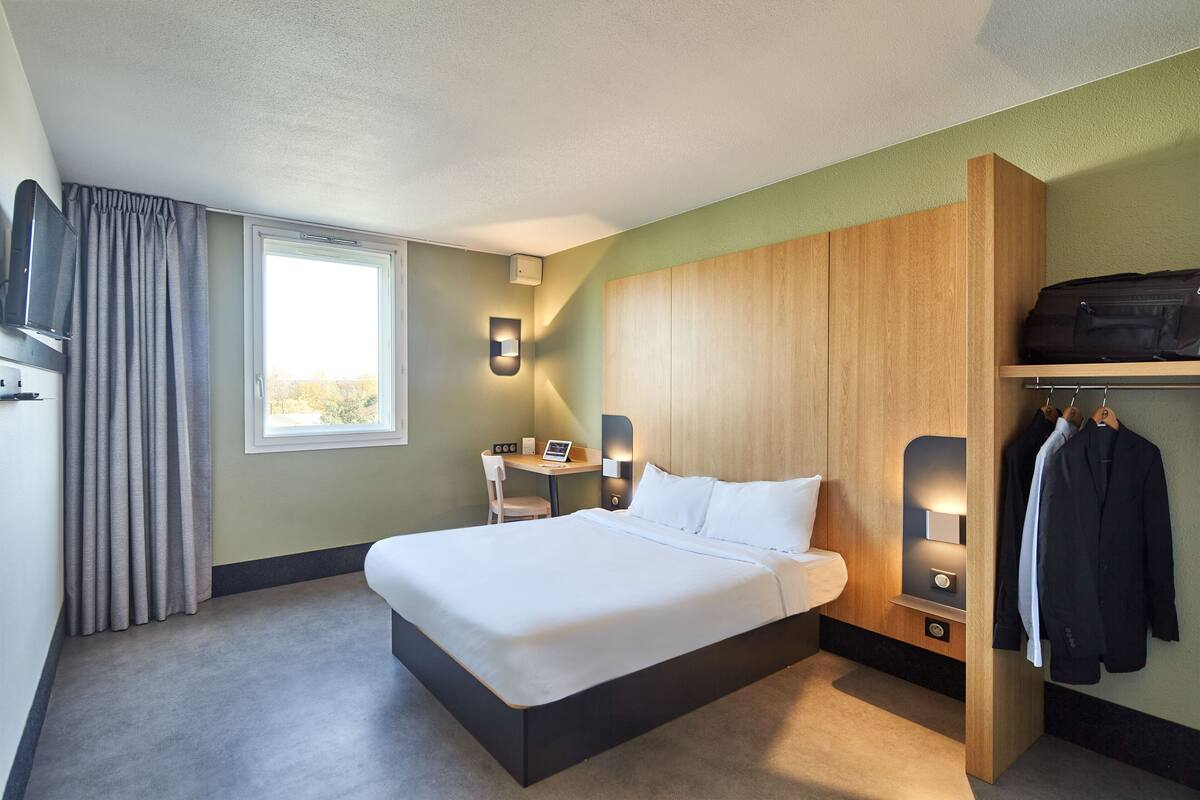 A hotel room with a large bed, a desk with a chair, a TV, and a coat rack with jackets.