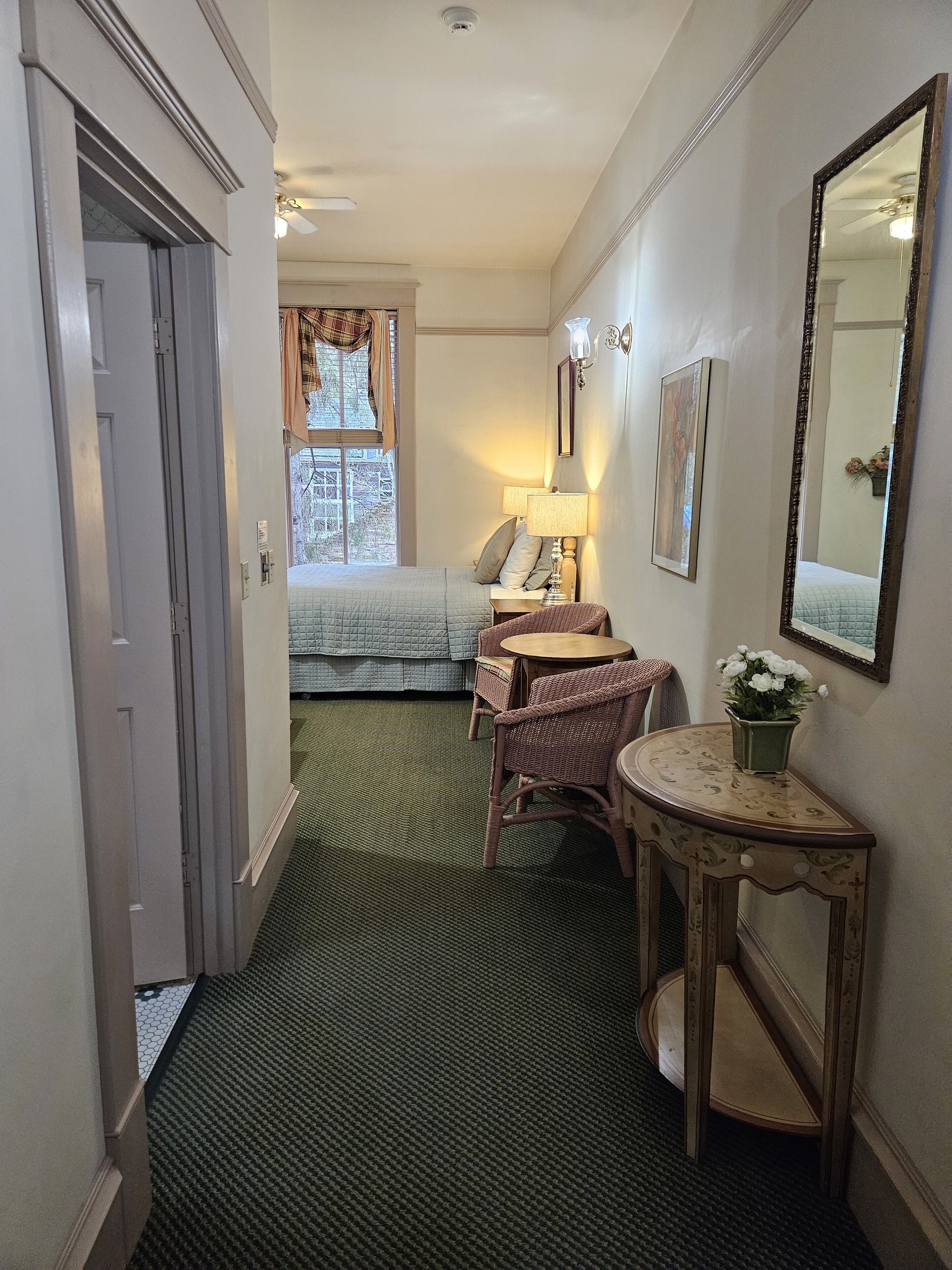 A narrow hallway leading to a bedroom with a bed, a nightstand, a lamp, and a small table with a vase of flowers.