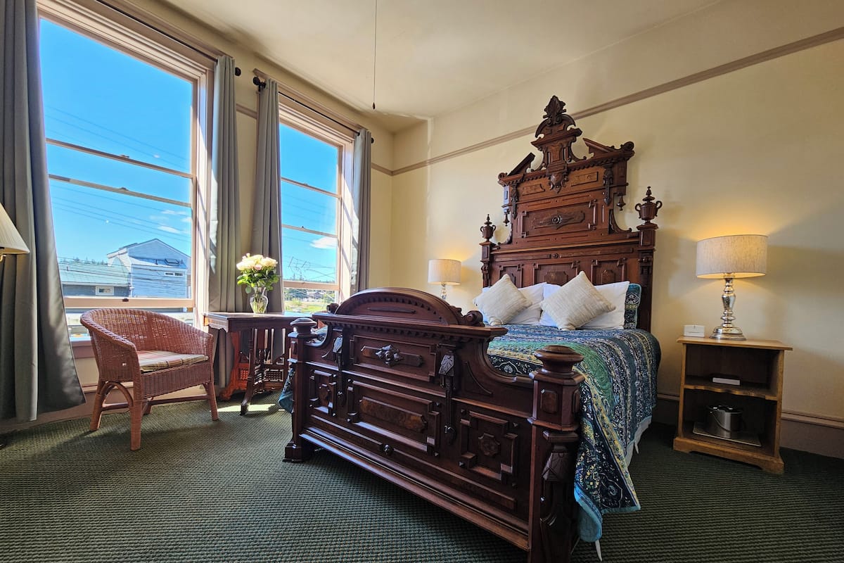A traditional wooden bed with a headboard, a wicker chair, a bedside table with a lamp, and a view of the outside through large windows.