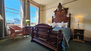 A traditional wooden bed with a headboard, a wicker chair, a bedside table with a lamp, and a view of the outside through large windows.