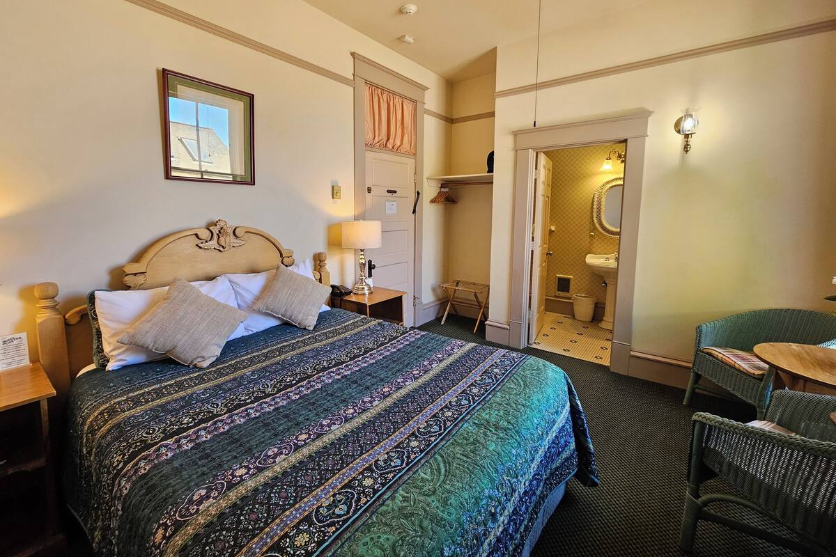 A hotel room with a large bed, a bedside table with a lamp, a chair, a small table, and a bathroom visible through an open door.