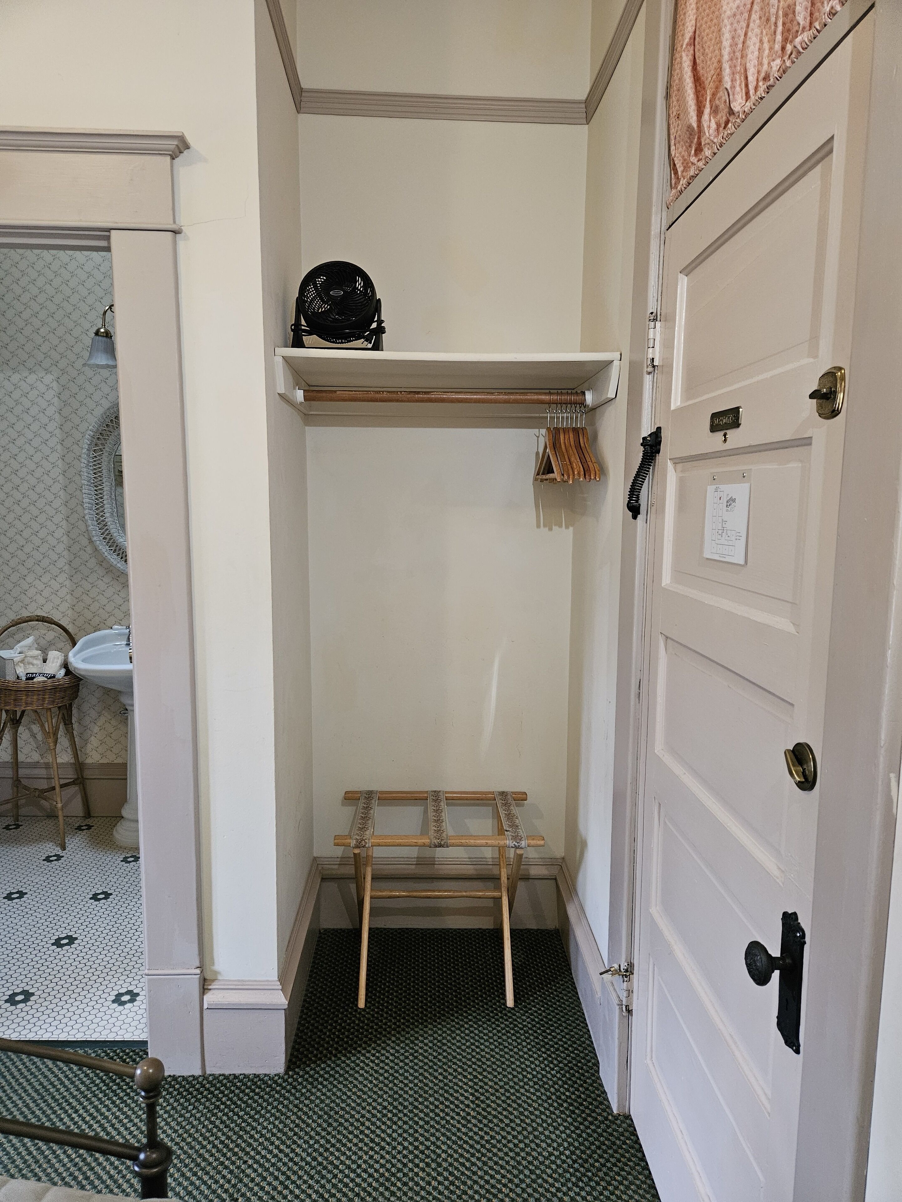 A corner room with a white door, a small shelf with a fan, a drying rack, and a patterned floor.