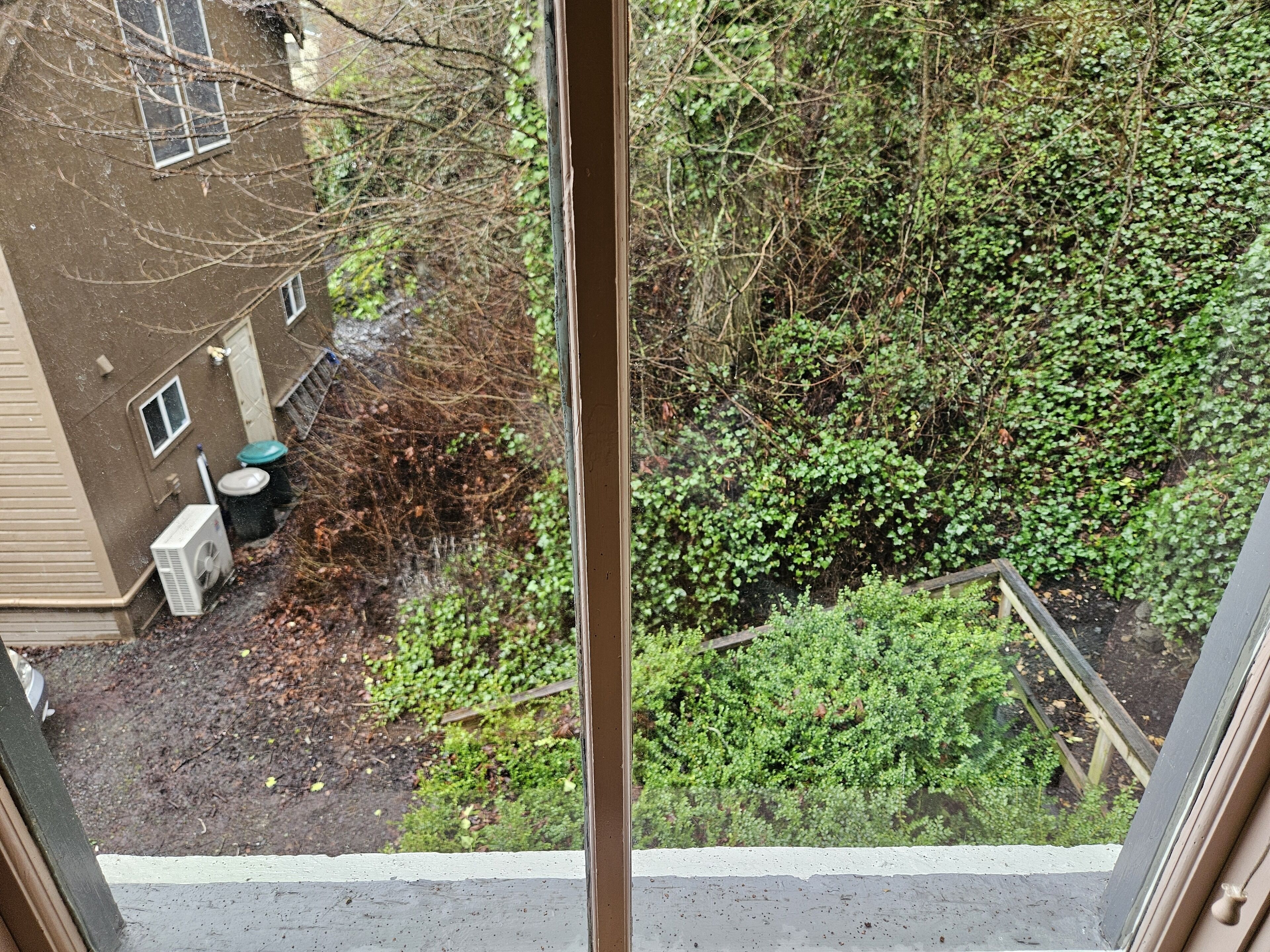 A view from a window overlooking a backyard with a brown building, a white air conditioning unit, and a green bush.