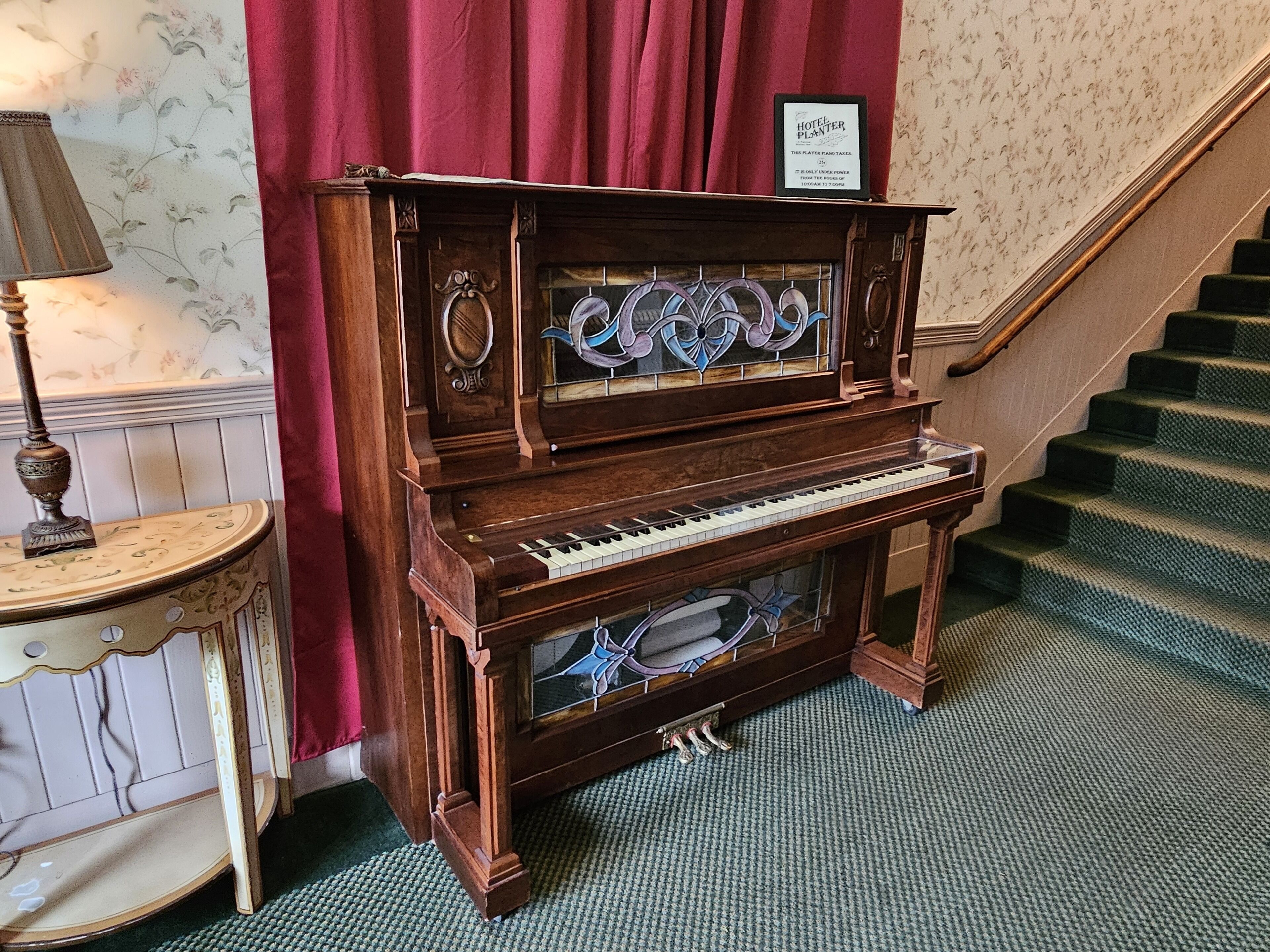Un piano de cola en el vestíbulo de un hotel, con una escalera y un cartel enmarcado encima.