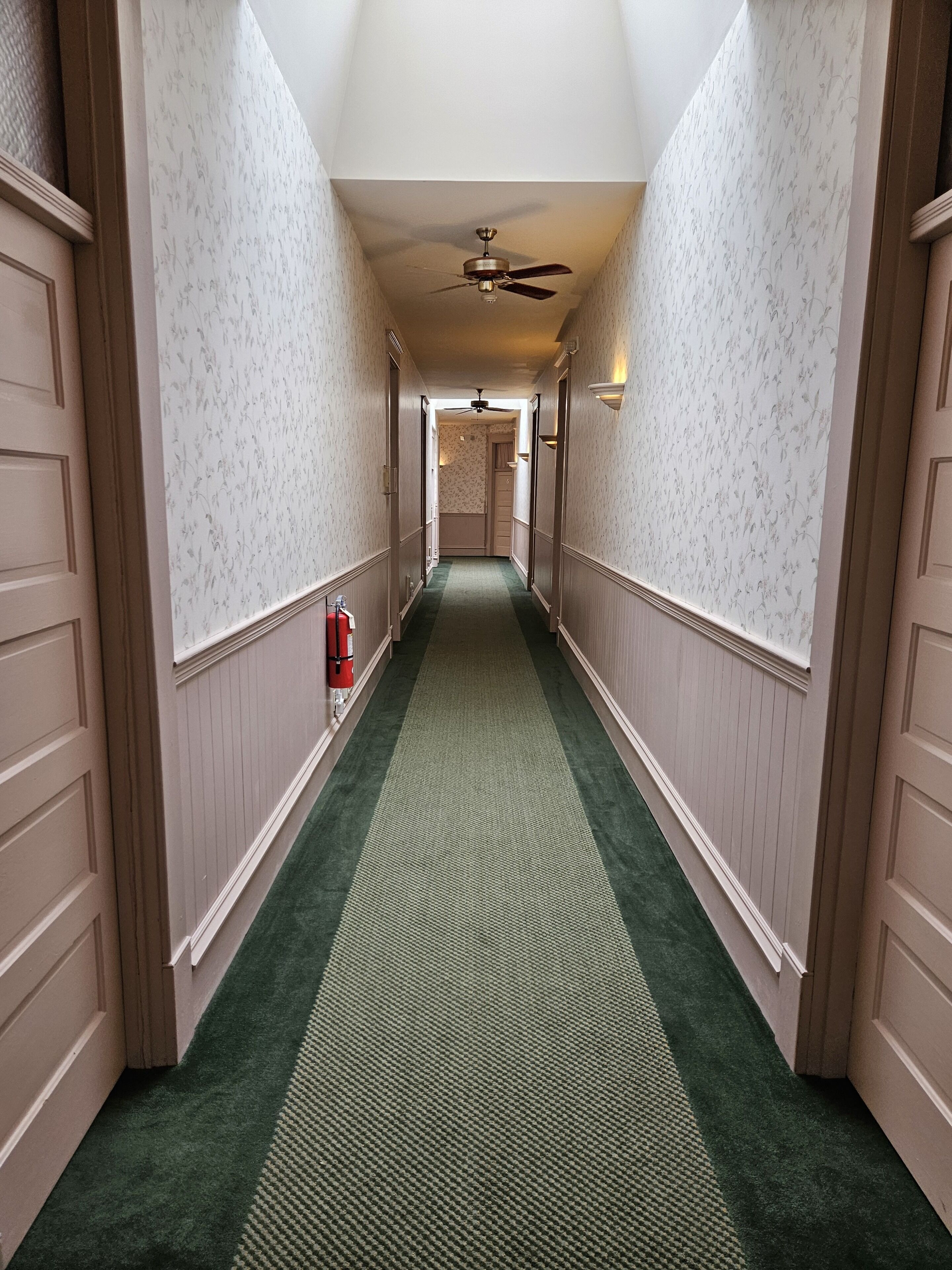A narrow hallway with patterned wallpaper, a green carpet, and a ceiling fan.